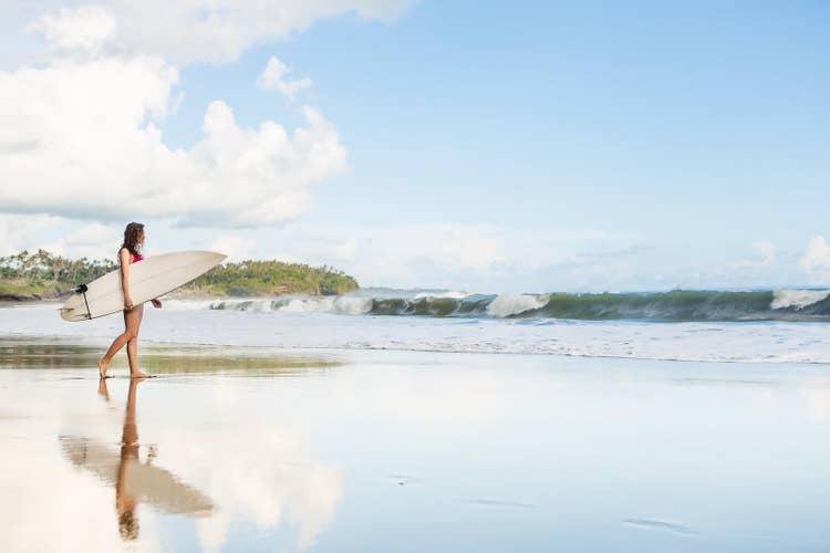 A surfer heading into the water at a coastal break — the same impulse that draws every wild swimmer toward open water: not the pool, not the lane rope, but the actual thing